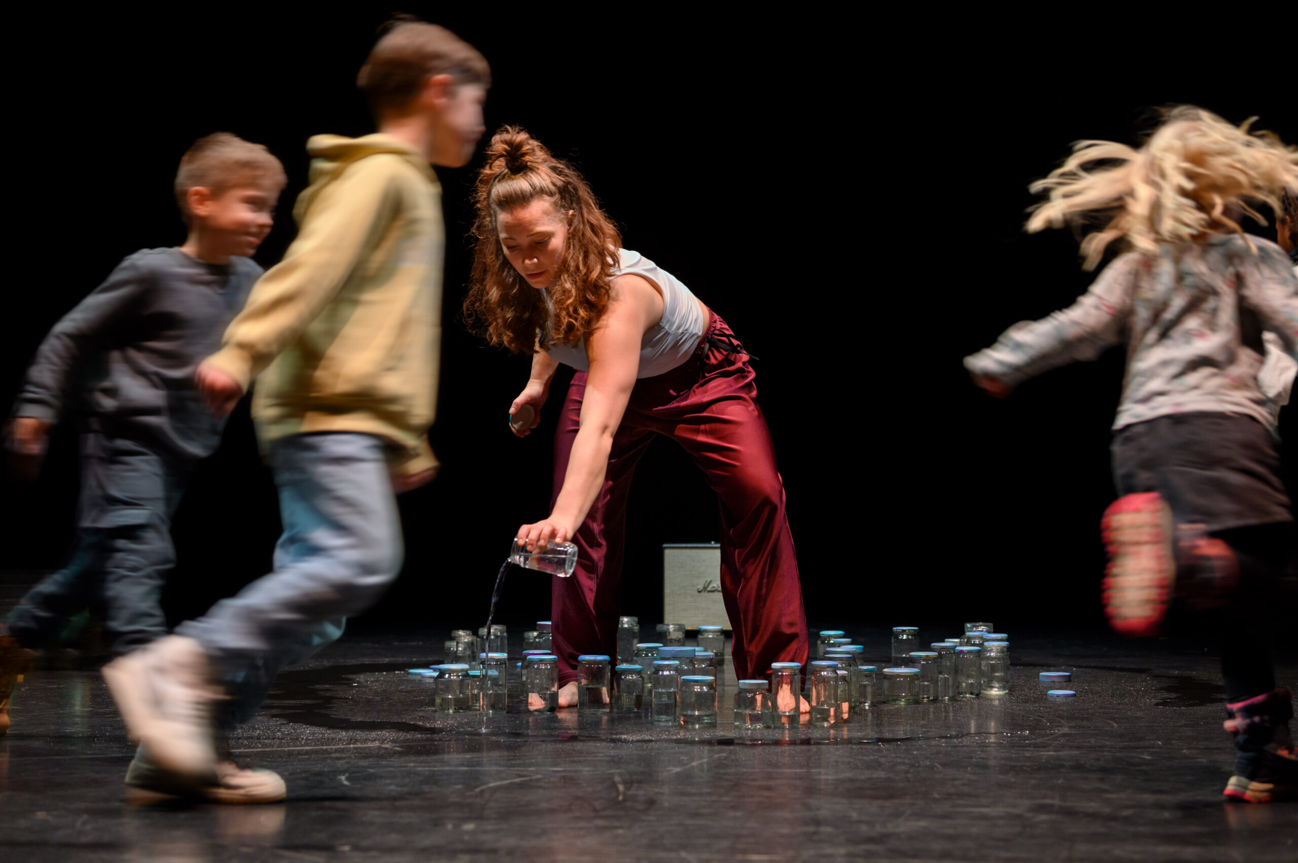 Kinderen rennen rond op podium, rondom glazen potjes. 